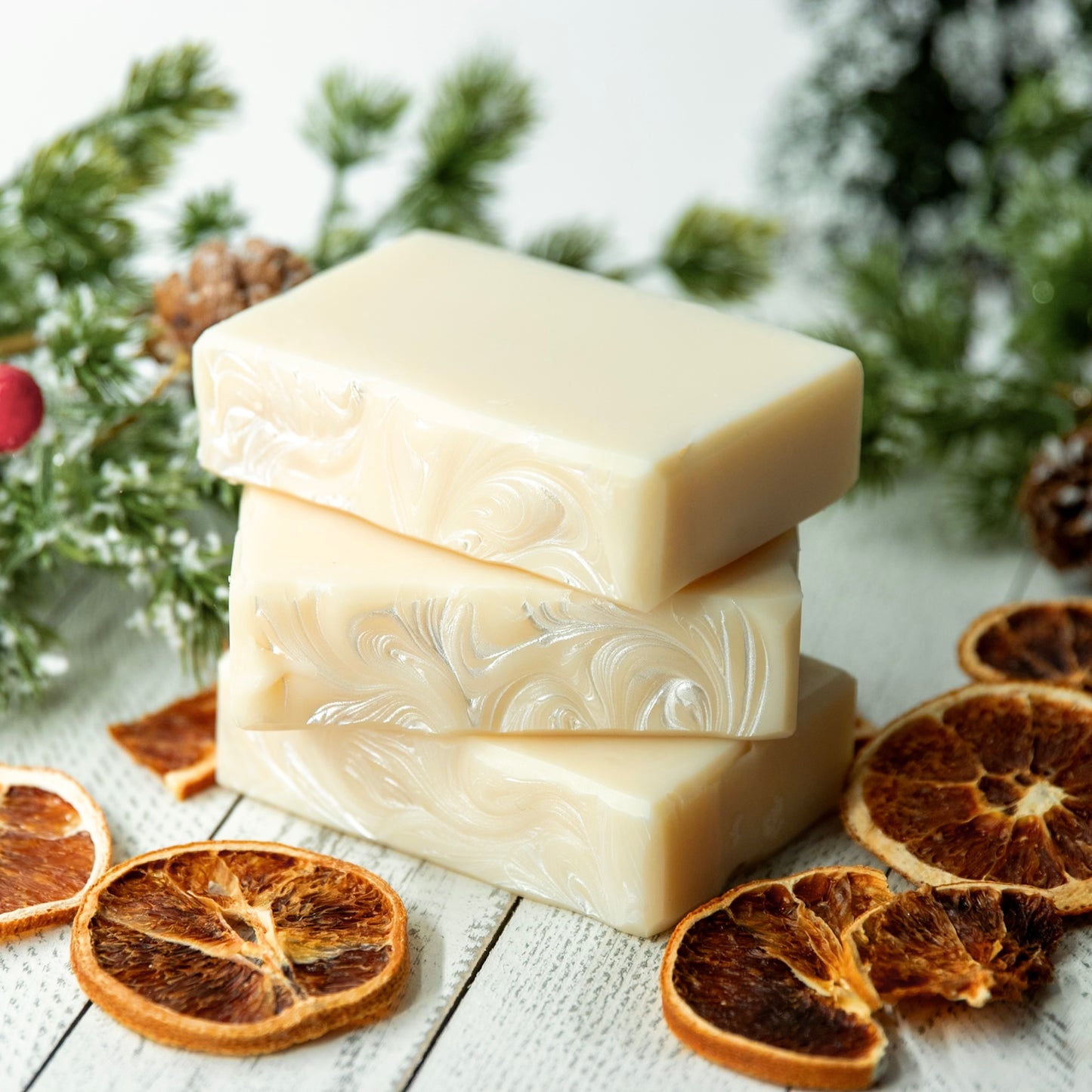 Two bars of soap stacked on a wooden surface with dried oranges and pine branches in the background.