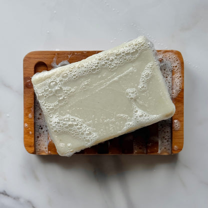 A rectangular bar of creamy off-white soap resting on a wooden soap dish, covered in lathered foam, set on a light marble surface.