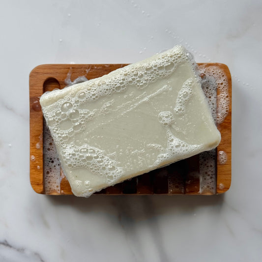 A rectangular bar of creamy off-white soap resting on a wooden soap dish, covered in lathered foam, set on a light marble surface.