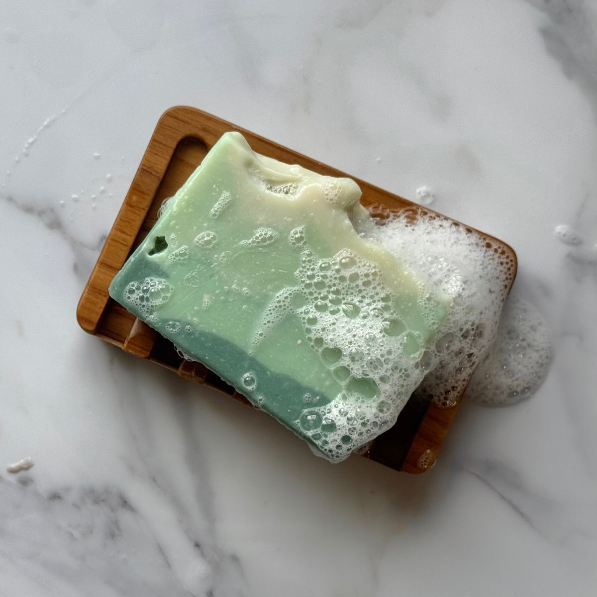 A rectangular bar of layered green and cream soap resting on a wooden soap dish, covered with white foam, on a white marble surface.