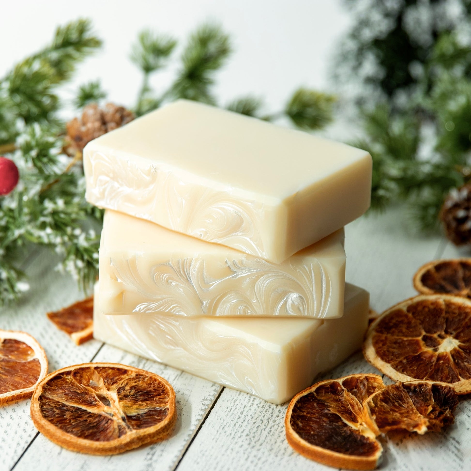 Two bars of soap stacked on a wooden surface with dried oranges and pine branches in the background.