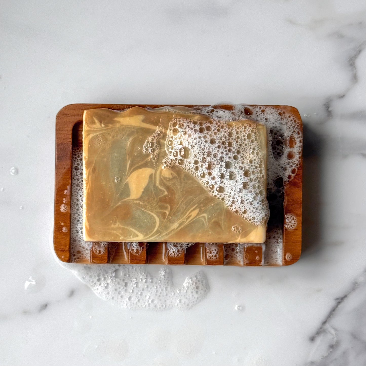 A rectangular bar of marbled golden-beige soap resting on a wooden soap dish filled with lather, placed on a white marble surface.