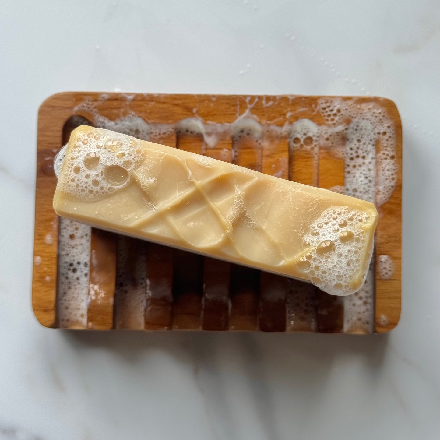 A rectangular bar of pale yellow soap with bubbly foam ends rests diagonally on a wooden soap dish over a white marble surface.