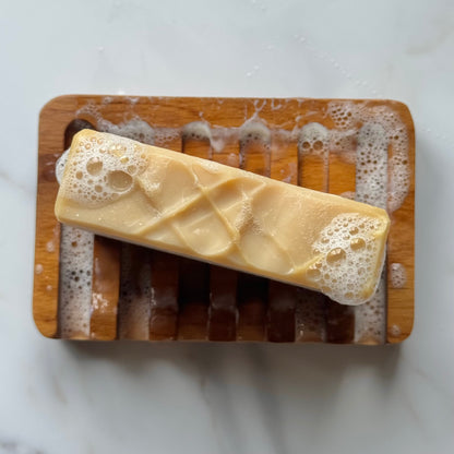 A rectangular bar of pale yellow soap with bubbly foam ends rests diagonally on a wooden soap dish over a white marble surface.